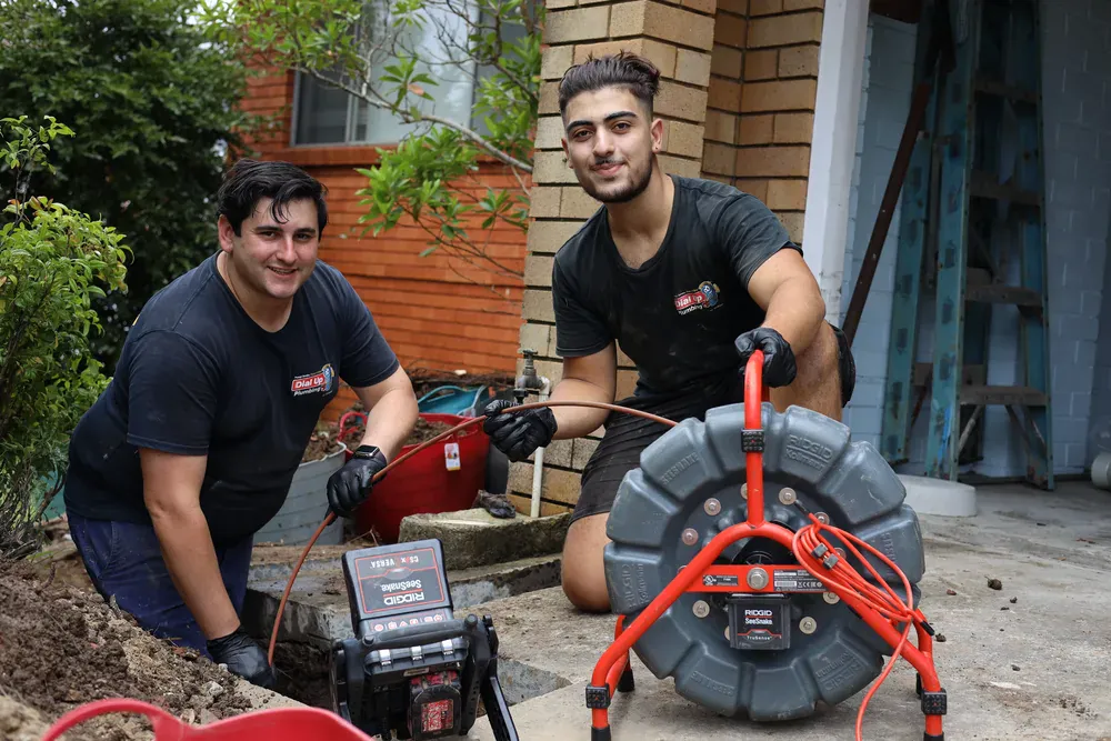 Dial Up plumbers running a drain camera reel beside a home